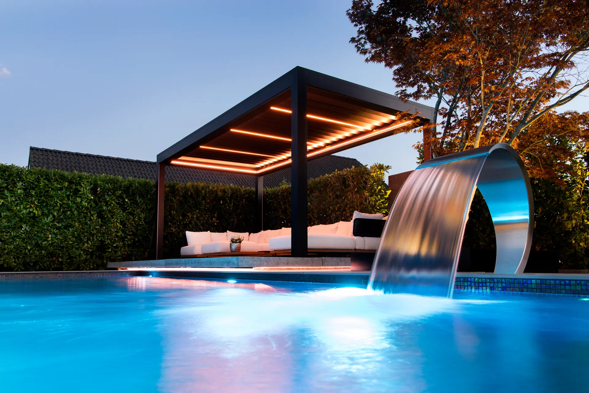 Brustor pergola with integrated warm LED lighting creating a cozy lounge area beside a modern swimming pool