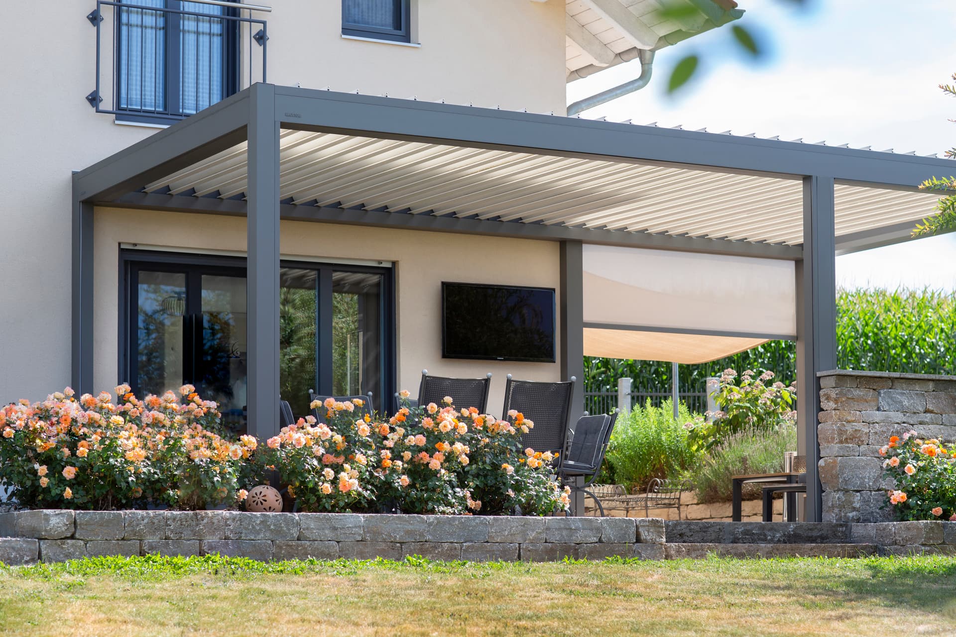 Aluminium pergola with a louvred roof installed on a terrace beside a house, shading an outdoor seating area surrounded by flowering plants.